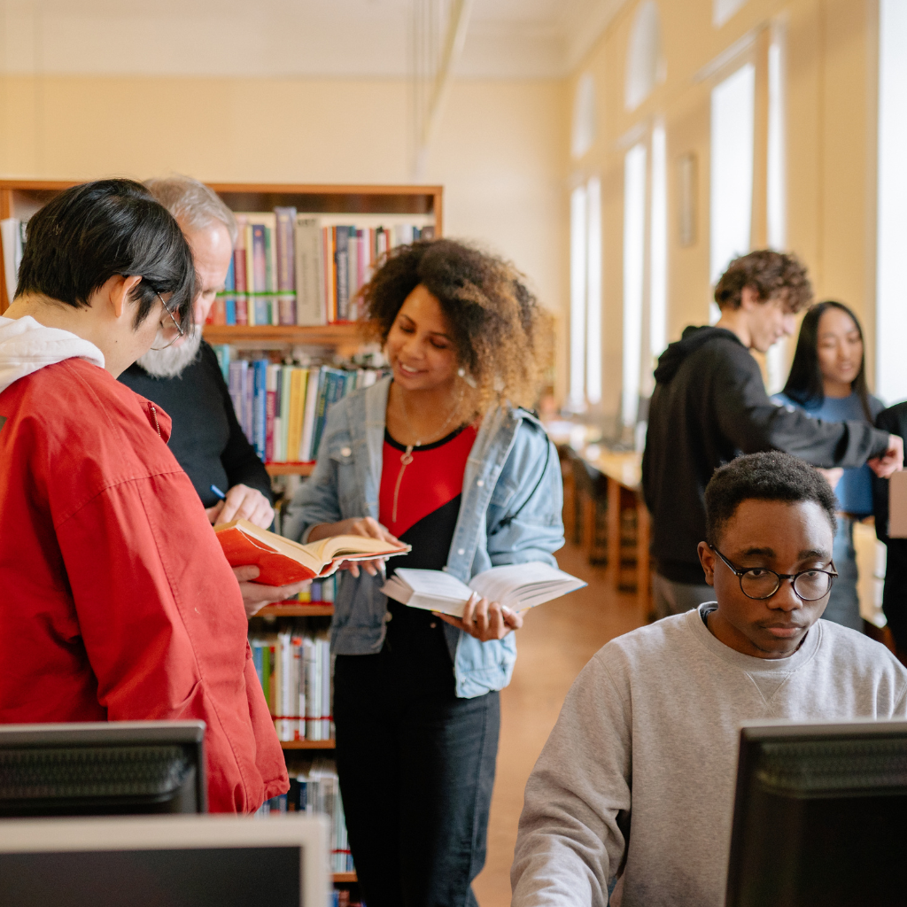 A group of people utilizing various services in a library, using books and a computer